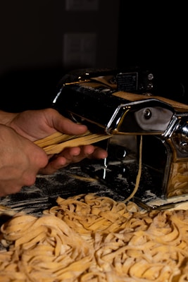 Hands are guiding freshly cut pasta coming out of a pasta machine, with a pile of flour-dusted pasta strands below. The machine has a shiny metal surface, reflecting the warm lighting in the room.