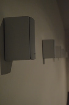 A series of rectangular speakers are mounted on a wall in a row. The lighting is dim, casting faint shadows on the wall. The foreground shows one speaker in clear focus, while the others fade into the background.