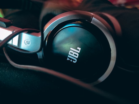 Close-up view of a pair of JBL headphones. The focus is on the ear cup which prominently features the JBL logo. The headphones appear to be in a dark setting with some ambient light reflecting off the surfaces.