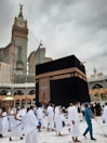 Pilgrims dressed in white garments are walking around the Kaaba, a cube-shaped structure in the center of a large mosque. A tall clock tower is visible in the background beneath a cloudy sky.
