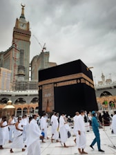 Pilgrims dressed in white garments are walking around the Kaaba, a cube-shaped structure in the center of a large mosque. A tall clock tower is visible in the background beneath a cloudy sky.