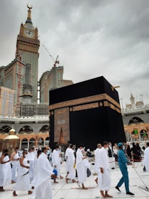 Pilgrims dressed in white garments are walking around the Kaaba, a cube-shaped structure in the center of a large mosque. A tall clock tower is visible in the background beneath a cloudy sky.