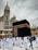 Pilgrims dressed in white garments are walking around the Kaaba, a cube-shaped structure in the center of a large mosque. A tall clock tower is visible in the background beneath a cloudy sky.