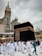 Pilgrims dressed in white garments are walking around the Kaaba, a cube-shaped structure in the center of a large mosque. A tall clock tower is visible in the background beneath a cloudy sky.