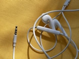 White earphones with a 3.5mm audio jack are placed on a textured yellow surface. The cable is loosely coiled, and the earbud design suggests they are typical in-ear headphones. There is a visible control button on the cable.