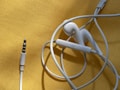 White earphones with a 3.5mm audio jack are placed on a textured yellow surface. The cable is loosely coiled, and the earbud design suggests they are typical in-ear headphones. There is a visible control button on the cable.