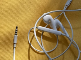 White earphones with a 3.5mm audio jack are placed on a textured yellow surface. The cable is loosely coiled, and the earbud design suggests they are typical in-ear headphones. There is a visible control button on the cable.