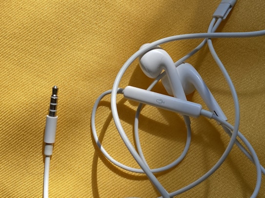 White earphones with a 3.5mm audio jack are placed on a textured yellow surface. The cable is loosely coiled, and the earbud design suggests they are typical in-ear headphones. There is a visible control button on the cable.