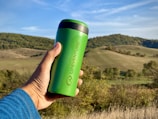 A green reusable travel mug is held by a hand in front of a picturesque landscape with rolling hills and blue sky.