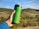 A green reusable travel mug is held by a hand in front of a picturesque landscape with rolling hills and blue sky.