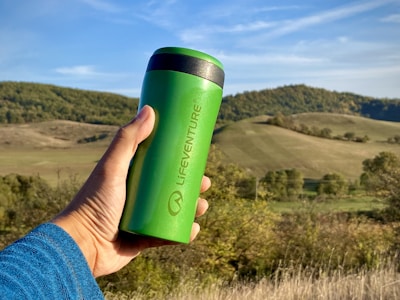 A green reusable travel mug is held by a hand in front of a picturesque landscape with rolling hills and blue sky.
