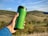 A green reusable travel mug is held by a hand in front of a picturesque landscape with rolling hills and blue sky.