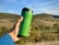 A green reusable travel mug is held by a hand in front of a picturesque landscape with rolling hills and blue sky.