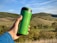 A green reusable travel mug is held by a hand in front of a picturesque landscape with rolling hills and blue sky.