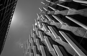 A black and white photograph of modern architecture, featuring a building façade with vertical, elongated panels casting dynamic shadows. The angle captures the structure looking upward towards the sky, with a second building visible on the left and a sunflare at the top left corner.