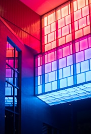 Colorful architectural interior featuring geometric window panels. The lighting casts vibrant hues of red, pink, and blue, creating a modern and artistic ambiance.