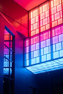 Colorful architectural interior featuring geometric window panels. The lighting casts vibrant hues of red, pink, and blue, creating a modern and artistic ambiance.
