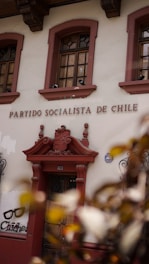 The image displays the facade of a building with the words 'Partido Socialista de Chile' above a wooden door. The building is adorned with ornate architectural details and flanked by two windows with intricate ironwork. There is also graffiti art on the wall nearby featuring a pair of glasses and the text 'Chicho'. Leaves partially obscure the lower corner of the image.