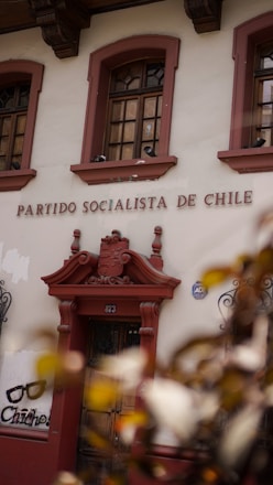The image displays the facade of a building with the words 'Partido Socialista de Chile' above a wooden door. The building is adorned with ornate architectural details and flanked by two windows with intricate ironwork. There is also graffiti art on the wall nearby featuring a pair of glasses and the text 'Chicho'. Leaves partially obscure the lower corner of the image.