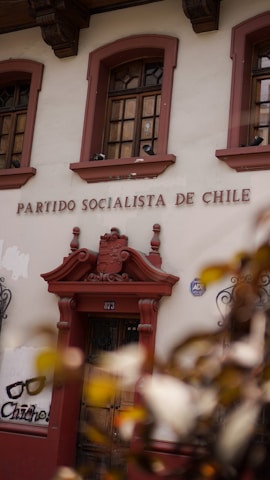 The image displays the facade of a building with the words 'Partido Socialista de Chile' above a wooden door. The building is adorned with ornate architectural details and flanked by two windows with intricate ironwork. There is also graffiti art on the wall nearby featuring a pair of glasses and the text 'Chicho'. Leaves partially obscure the lower corner of the image.