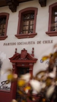 The image displays the facade of a building with the words 'Partido Socialista de Chile' above a wooden door. The building is adorned with ornate architectural details and flanked by two windows with intricate ironwork. There is also graffiti art on the wall nearby featuring a pair of glasses and the text 'Chicho'. Leaves partially obscure the lower corner of the image.