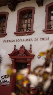 The image displays the facade of a building with the words 'Partido Socialista de Chile' above a wooden door. The building is adorned with ornate architectural details and flanked by two windows with intricate ironwork. There is also graffiti art on the wall nearby featuring a pair of glasses and the text 'Chicho'. Leaves partially obscure the lower corner of the image.