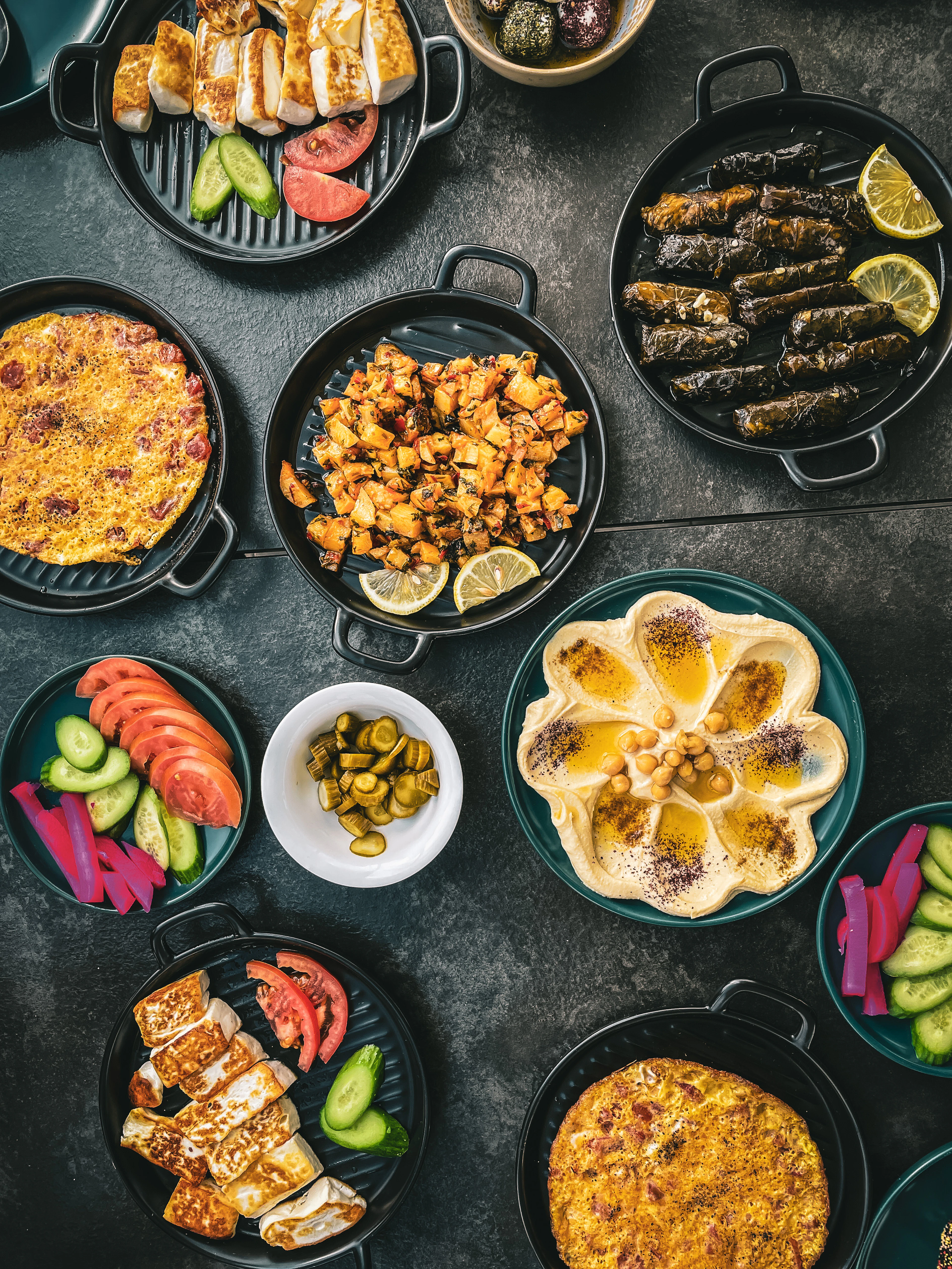 A Levantine mezze spread photographed in Amman, Jordan - grilled halloumi, stuffed vine leaves, hummus and foul pooled with olive oil, spiced potatoes, pickles, tomato salad and pink pickled turnips served in small black pans