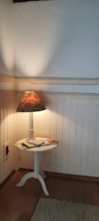 A small round white wooden table with a floral-patterned lamp on top, positioned in the corner of a room with white paneled walls. The table holds a few items, including a notepad and what appears to be a pen. The warm glow of the lamp creates a cozy atmosphere.