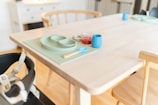 A wooden dining table is set with a green silicone placemat, a matching divided plate, and a small blue cup. Beside these, there is a glass jar filled with a red substance, possibly jam. A child’s high chair with safety straps is positioned next to the table.