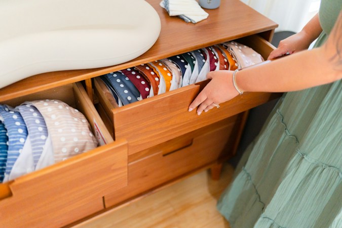 Parent tucking folded cloth diapers into an open wooden dresser drawer below a changing pad
