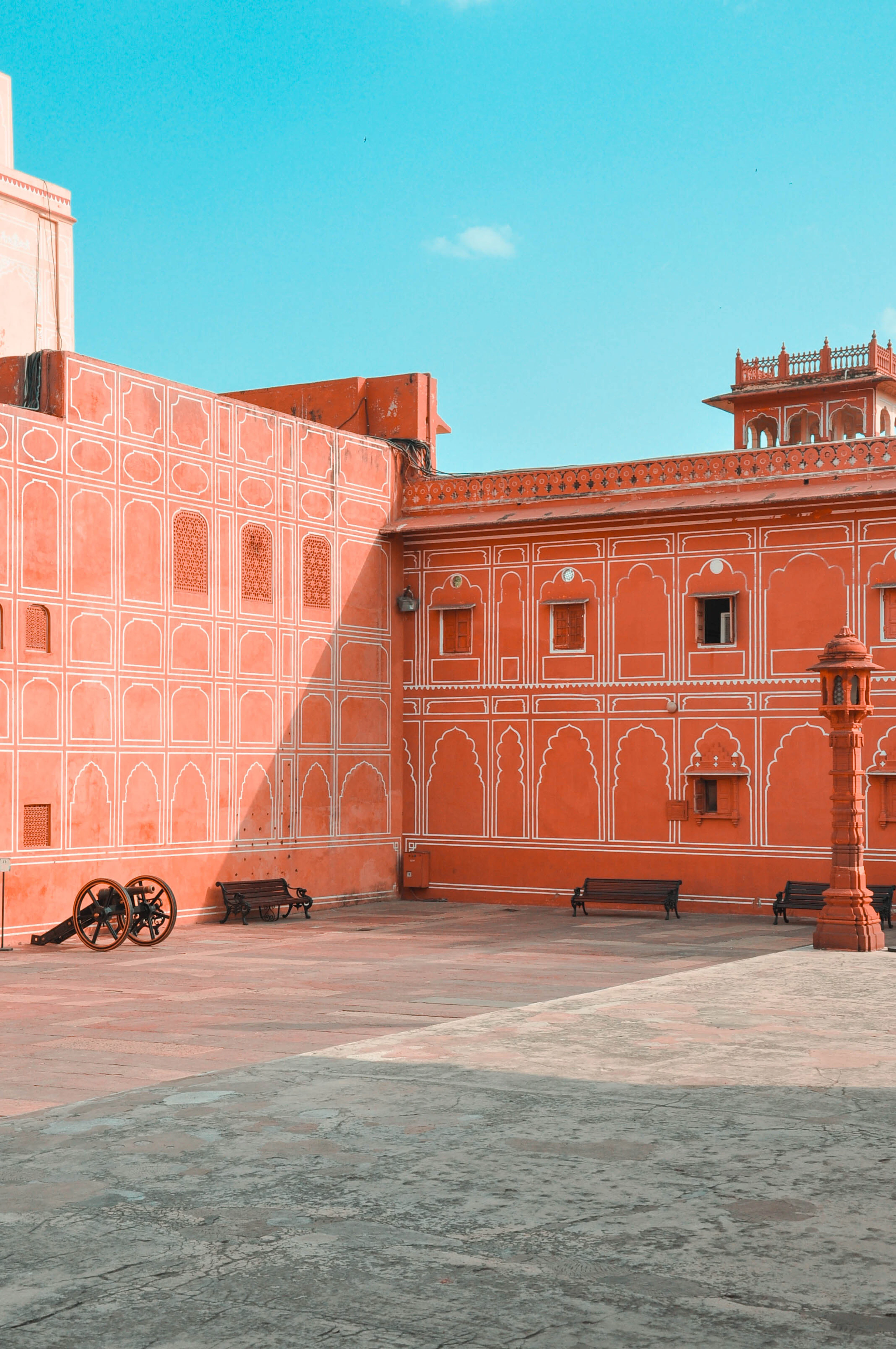 A terracotta-pink Jaipur courtyard wall with white stencil patterns, an old cannon, and empty benches.