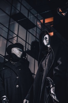 Two mannequins are dressed in dark, dramatic clothing. The one on the left wears a wide-brimmed hat and coat with buttons, while the one on the right is draped in a cloak with a hood. The background features a grid of reflective panels with some warm colored light seeping through.