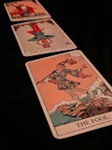 Three tarot cards are laid out diagonally. The closest card is 'The Fool,' depicting a figure in colorful attire holding a stick with a bag and a flower, standing on the edge of a cliff. The middle card is 'The Hanged Man,' showing a figure hanging upside down by one foot. The furthest card is 'The Magician,' displaying a figure in a red robe gesturing with one hand up and one hand down.