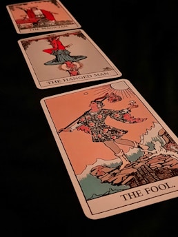 Three tarot cards are laid out diagonally. The closest card is 'The Fool,' depicting a figure in colorful attire holding a stick with a bag and a flower, standing on the edge of a cliff. The middle card is 'The Hanged Man,' showing a figure hanging upside down by one foot. The furthest card is 'The Magician,' displaying a figure in a red robe gesturing with one hand up and one hand down.