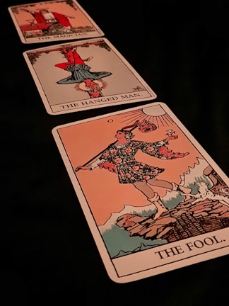 Three tarot cards are laid out diagonally. The closest card is 'The Fool,' depicting a figure in colorful attire holding a stick with a bag and a flower, standing on the edge of a cliff. The middle card is 'The Hanged Man,' showing a figure hanging upside down by one foot. The furthest card is 'The Magician,' displaying a figure in a red robe gesturing with one hand up and one hand down.