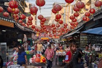 The image depicts a bustling street market lined with numerous red Chinese lanterns hanging above. The market is crowded with people moving about, some browsing various stalls that sell a range of goods. Buildings flanking the street have signage in an Asian script. The vibrant atmosphere suggests a lively market day with a cultural festival or celebration.