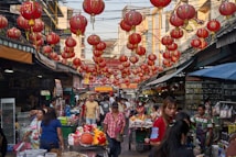 The image depicts a bustling street market lined with numerous red Chinese lanterns hanging above. The market is crowded with people moving about, some browsing various stalls that sell a range of goods. Buildings flanking the street have signage in an Asian script. The vibrant atmosphere suggests a lively market day with a cultural festival or celebration.