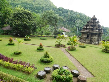 A lush green garden featuring manicured lawns and trimmed shrubs, with a stone pathway leading to an ancient temple structure. The background is dominated by a dense forest and a small pavilion is visible among the trees.