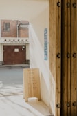 A wooden signboard displaying opening hours and floor information stands on a sunlit floor next to a wall, leading to a view of an alley with a brick building and a closed door in the background. Posters are affixed to the wall beside the sign.