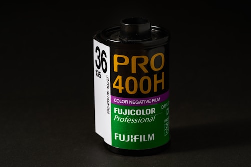 A roll of Fujifilm Pro 400H color negative film is displayed prominently. The cylindrical film canister is upright with a label indicating it is a professional-grade film with 36 exposures. The label features white, purple, and green colors, with bold yellow lettering for the film type and the Fujifilm logo.