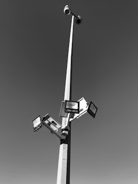 A tall pole with multiple solar-powered lights and surveillance cameras extending outward against a clear sky. The pole is robust and slender, creating a geometric structure with clean lines.