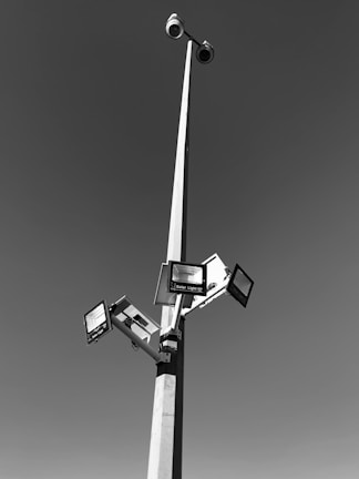 A tall pole with multiple solar-powered lights and surveillance cameras extending outward against a clear sky. The pole is robust and slender, creating a geometric structure with clean lines.
