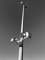 A tall pole with multiple solar-powered lights and surveillance cameras extending outward against a clear sky. The pole is robust and slender, creating a geometric structure with clean lines.