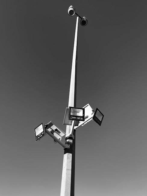 A tall pole with multiple solar-powered lights and surveillance cameras extending outward against a clear sky. The pole is robust and slender, creating a geometric structure with clean lines.