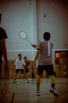 Several people are engaged in a game of badminton indoors, with one player at the forefront holding a racket. The scene captures the dynamic action and focus of the participants, while cardboard boxes can be seen in the background, indicating the setting might be a multi-purpose indoor space.