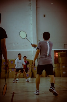 Several people are engaged in a game of badminton indoors, with one player at the forefront holding a racket. The scene captures the dynamic action and focus of the participants, while cardboard boxes can be seen in the background, indicating the setting might be a multi-purpose indoor space.