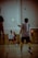 Several people are engaged in a game of badminton indoors, with one player at the forefront holding a racket. The scene captures the dynamic action and focus of the participants, while cardboard boxes can be seen in the background, indicating the setting might be a multi-purpose indoor space.