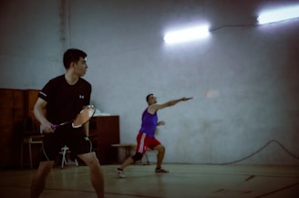 Two men are playing badminton indoors. One player is preparing to hit the shuttlecock, while the other is in a ready stance waiting for the play. The players are dressed in sports attire, and the lighting is dim with fluorescent lights overhead.