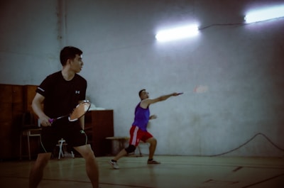 Two men are playing badminton indoors. One player is preparing to hit the shuttlecock, while the other is in a ready stance waiting for the play. The players are dressed in sports attire, and the lighting is dim with fluorescent lights overhead.