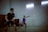 Two men are playing badminton indoors. One player is preparing to hit the shuttlecock, while the other is in a ready stance waiting for the play. The players are dressed in sports attire, and the lighting is dim with fluorescent lights overhead.